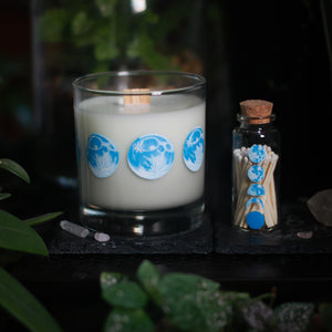 A white-wax candle with a wooden wick rests upon a dark surface with plants and crystals visible in the scene. To the right of it rests a 2.75" tall glass bottle, just a touch shorter than the candle. The candle glass has 8 phases of the moon painted in shades of blue circling the glass. The bottle has 5 phases of the moon painted in blue, painted vertically on the front of the bottle. The bottle is filled with wooden matches with white tops.