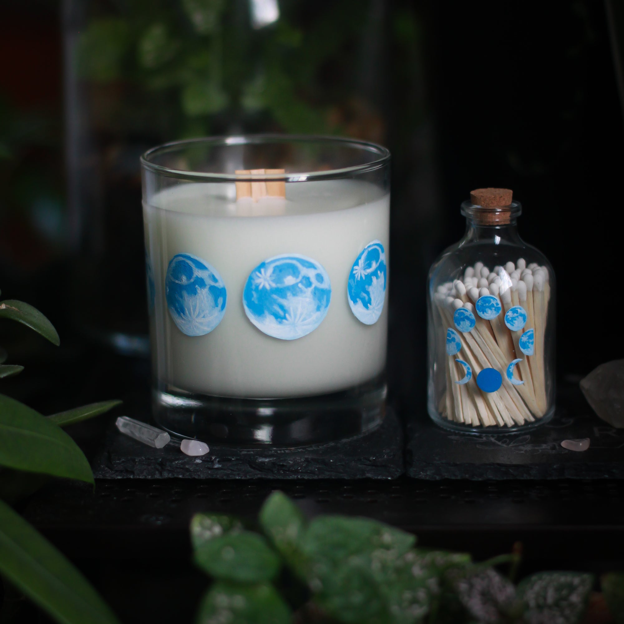 A white candle in a tumbler glass and with a wooden wick rests upon a dark surface with plants and crystals visible in the scene. To the right of it rests a 3" tall glass bottle with a cork top, just a touch shorter than the candle. The candle glass has 8 phases of the moon painted in blue circling the glass. The bottle also has 8 phases of the moon painted in blue, all arranged in a circle and visible on the front of the bottle. The bottle is filled with wooden matches with white tops.