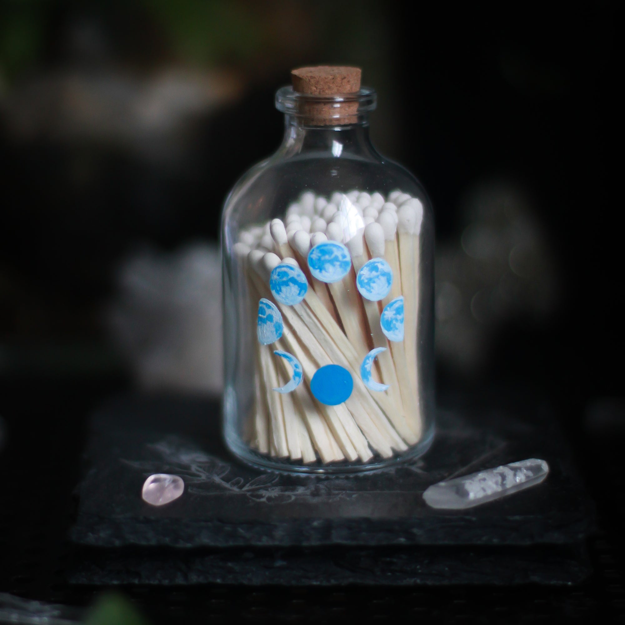 A glass bottle, 3" tall and 1.5" in diameter, with a cork top rests upon a dark surface and background with some crystals visible in the scene. The bottle has 8 phases of the moon painted in shades of blue, all arranged in a circle and visible on the front of the bottle. The full moon is at the top of the arrangement and the new moon, painted in solid blue, is at the bottom. The bottle is filled with wooden matches with white tops.