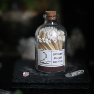 A glass bottle, 3" tall, 1.5" in diameter, with a cork top rests upon a dark surface and background with crystals in the scene. The bottle is filled with wooden matches with white tops. The back of the bottle is shown displaying a small white rectangular horizontal label. The label has gold lettering that reads "Jocelyn Arielle Designs" and "75 matches." There is also a gold crescent moon. Along the bottom of the label is a strip of striking paper to light the matches within it.