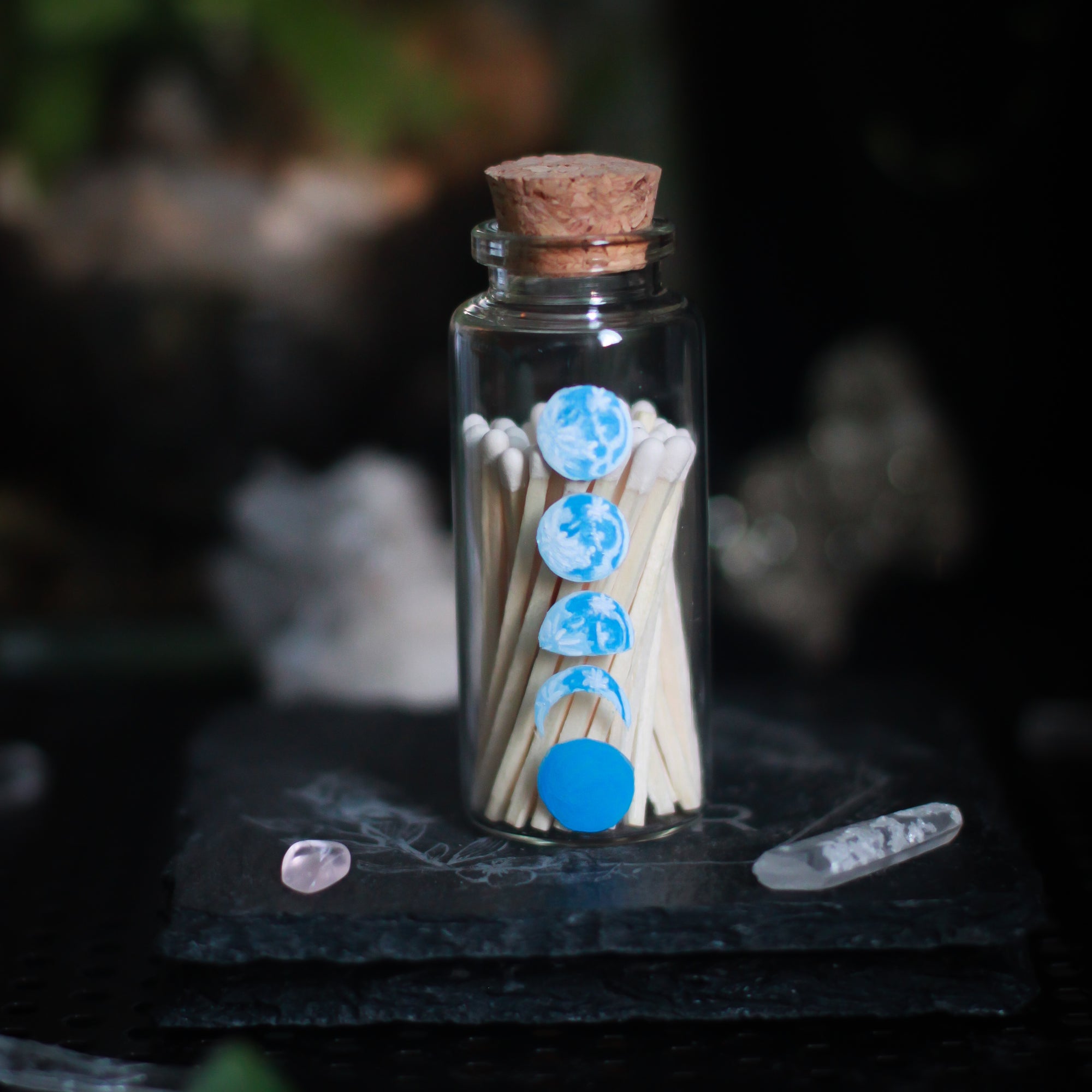 A small glass bottle, 2.75" tall and 1.18" in diameter, rests upon a dark surface and background with some crystals and plants visible in the scene. The bottle has 5 phases of the moon painted in blue, painted vertically on the front of the bottle. The bottle is filled with wooden matches with white tops.