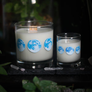 A white-wax filled candle in a tumbler glass and with a wooden wick rests upon a dark surface with plants and crystals visible in the scene. To the right of the tumbler candle is a smaller votive candle. Both candle glasses have 8 phases of the moon painted in shades of blue circling the glass. This angle depicts the full moons with two 3 quarter moons on either side of it