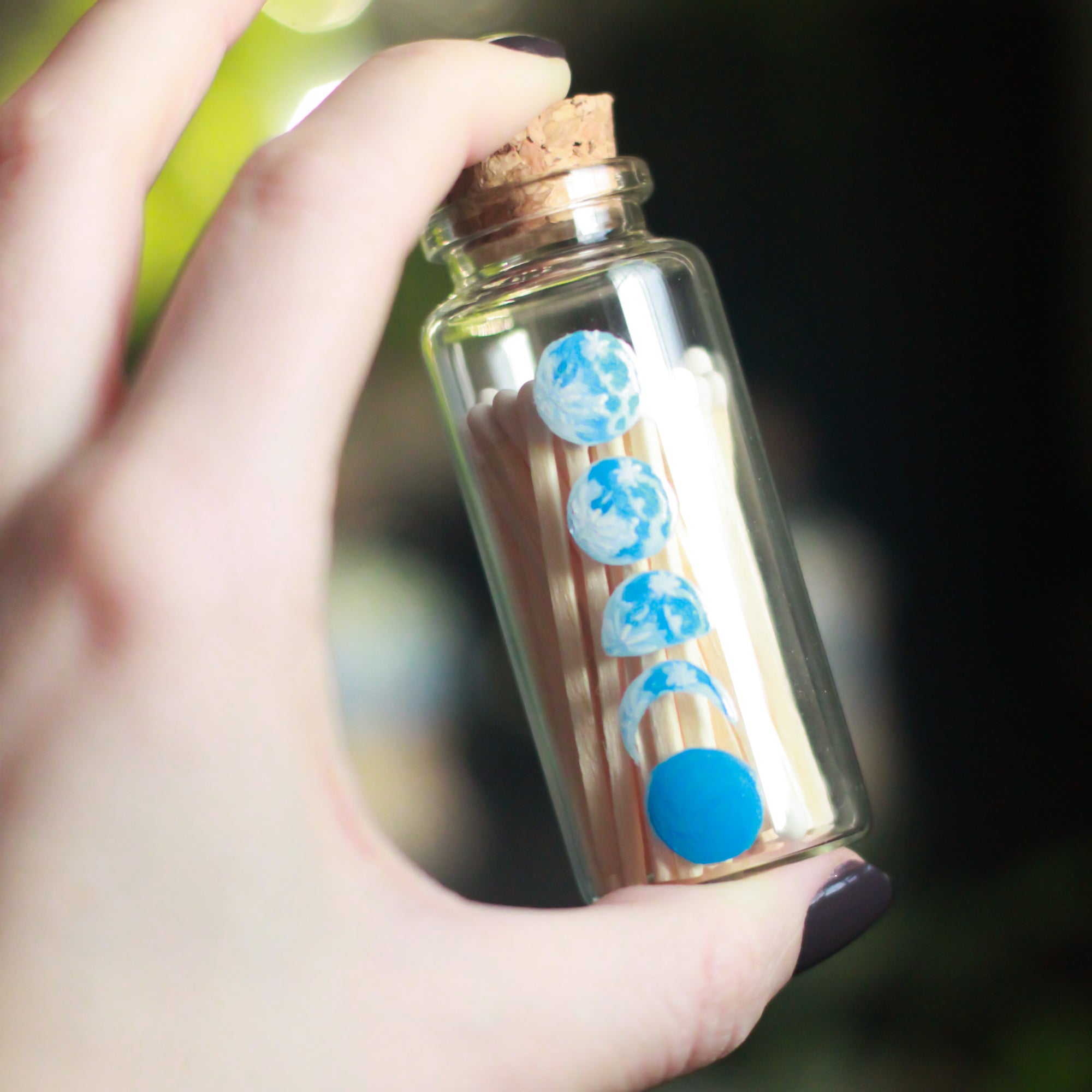 A small glass bottle, 2.75" tall and 1.18" in diameter, rests upon a dark surface and background with some crystals and plants visible in the scene. The bottle has 5 phases of the moon painted in blue, painted vertically on the front of the bottle. The bottle is filled with wooden matches with white tops.