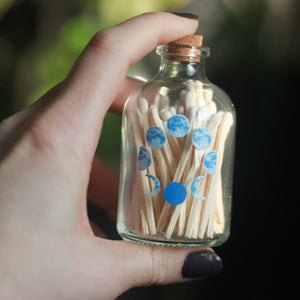 A small glass bottle, 3" tall and 1.5" in diameter, is held in a hand between the thumb and pointer finger. The bottle has 5 phases of the moon painted in blue, painted vertically on the front of the bottle. The bottle is filled with wooden matches with white tops.