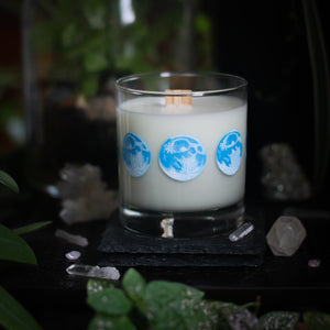 A white-wax candle with a wooden wick rests upon a dark surface with plants and crystals visible in the scene. The light candle stands out in contrast against the dark background. The candle glass has 8 phases of the moon painted in shades of blue circling the glass. This angle depicts the full moon with two 3 quarter moons on either side of it.