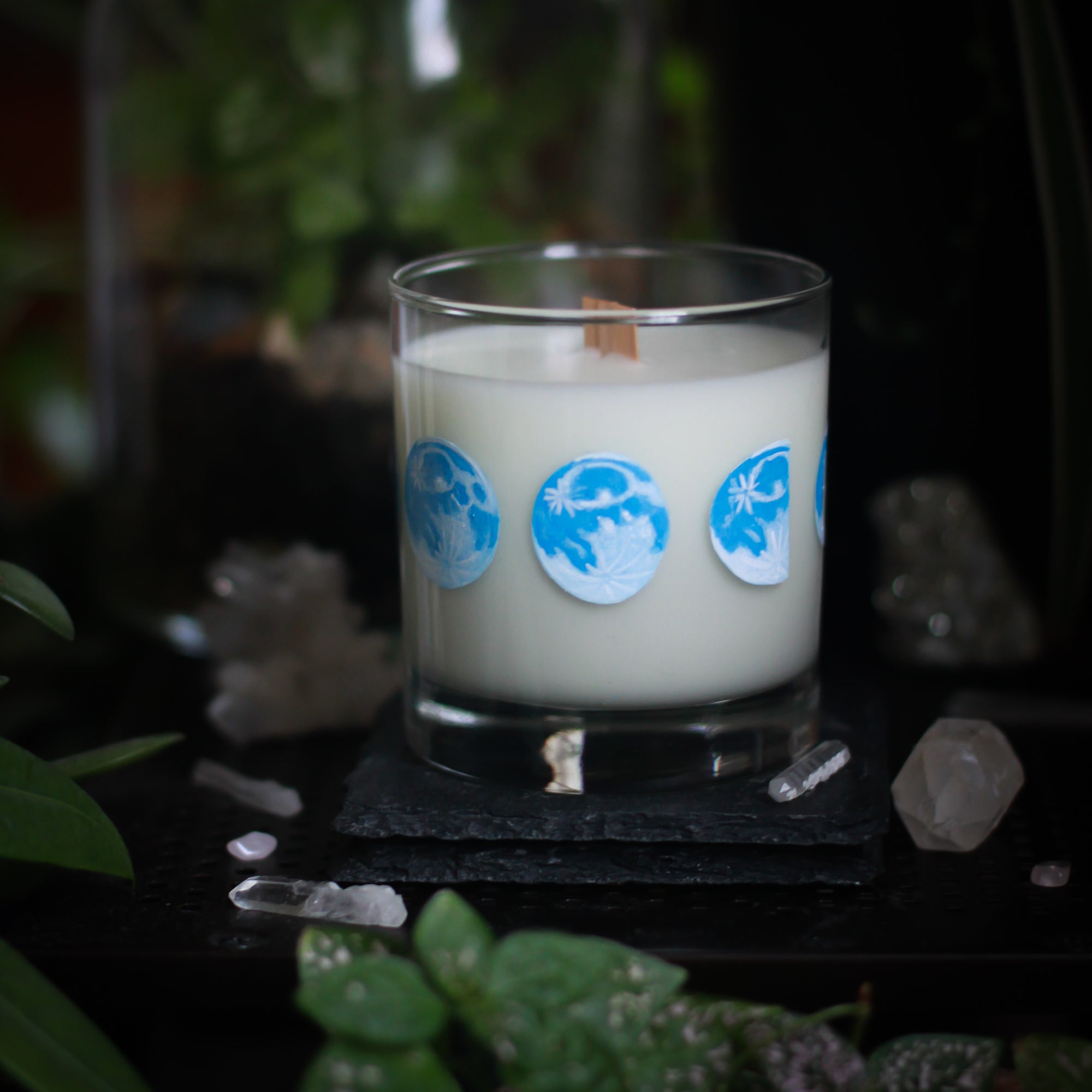 A white-wax candle with a wooden wick rests upon a dark surface with plants and crystals visible in the scene. The light candle stands out in contrast against the dark background. The candle glass has 8 phases of the moon painted in shades of blue circling the glass. This angle depicts the waning moon with the full moon to its left and a half moon to its right.