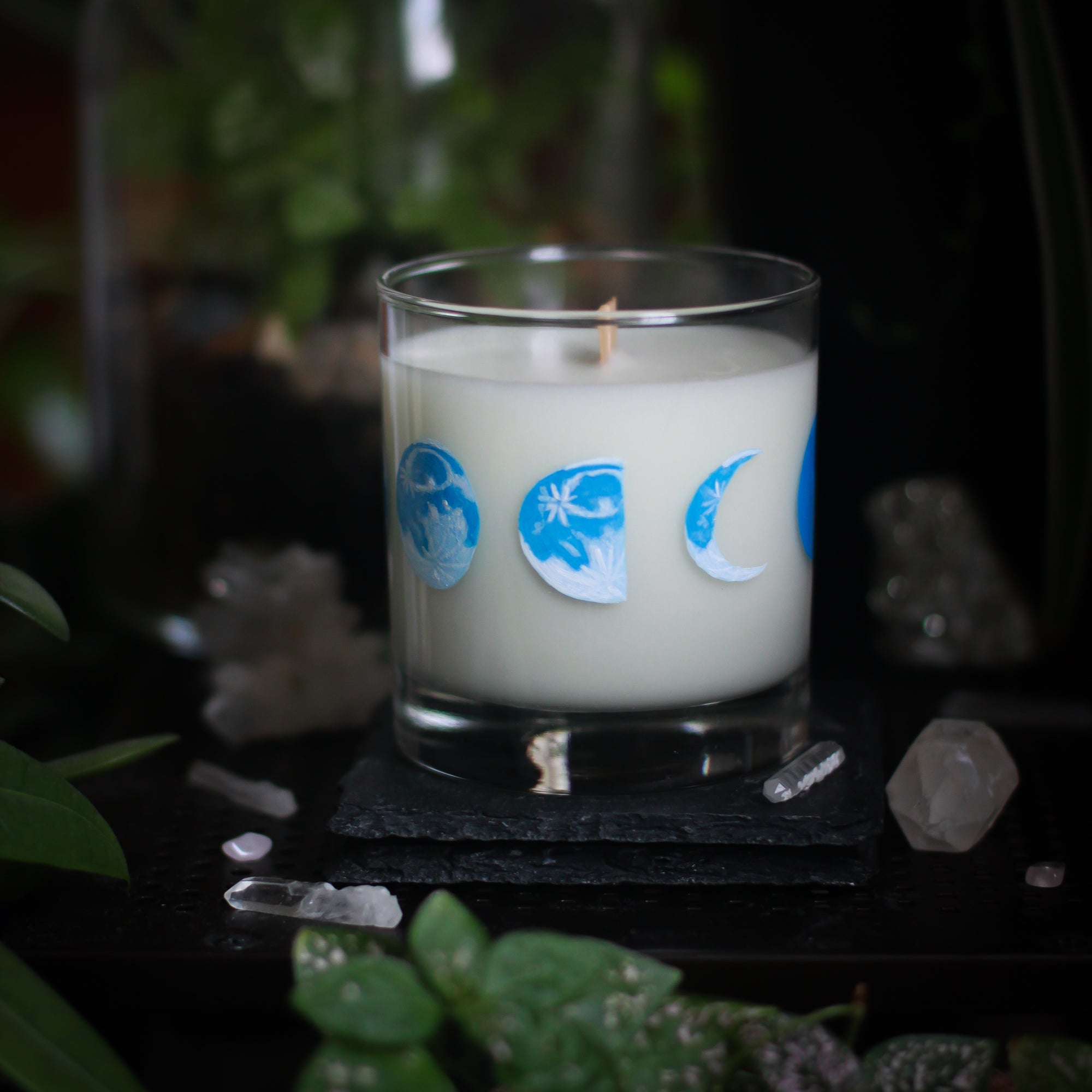 A white-wax candle with a wooden wick rests upon a dark surface with plants and crystals visible in the scene. The light candle stands out in contrast against the dark background. The candle glass has 8 phases of the moon painted in shades of blue circling the glass. This angle depicts the waning half moon centered with the waning gibbous moon to its left and the crescent moon to its right.