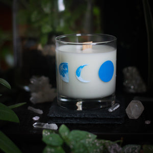 A white-wax candle with a wooden wick rests upon a dark surface with plants and crystals visible in the scene. The light candle stands out in contrast against the dark background. The candle glass has 8 phases of the moon painted in shades of blue circling the glass. This angle depicts the waning crescent moon at the center with the waning half moon to the left and the new moon, painting in solid blue, to the right.