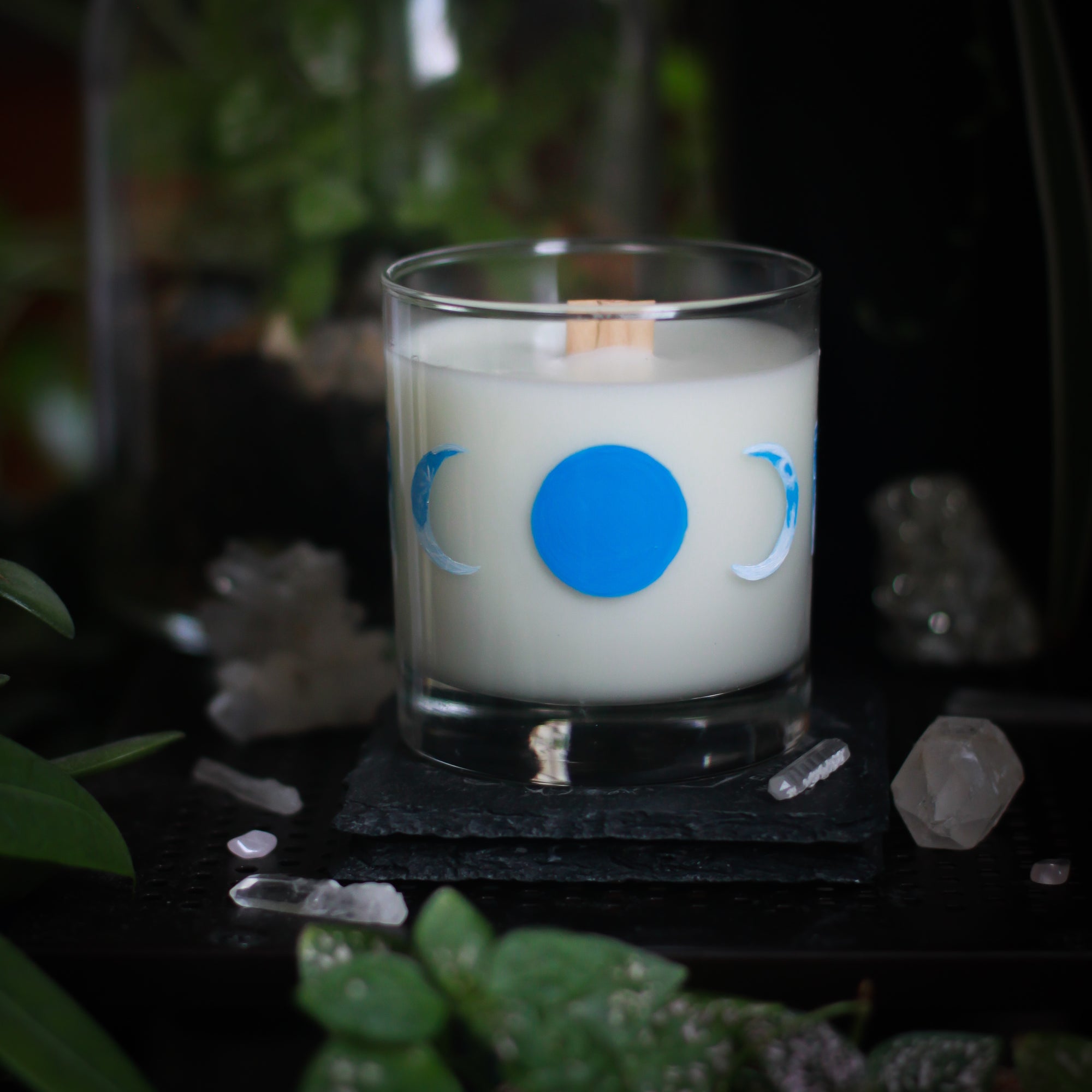 A white-wax candle with a wooden wick rests upon a dark surface with plants and crystals visible in the scene. The light candle stands out in contrast against the dark background. The candle glass has 8 phases of the moon painted in shades of blue circling the glass. This angle depicts the new moon, painted in solid blue, centered, with two crescent moons on either side facing inward towards it.