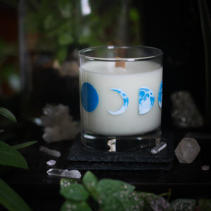 A white-wax candle with a wooden wick rests upon a dark surface with plants and crystals visible in the scene. The light candle stands out in contrast against the dark background. The candle glass has 8 phases of the moon painted in shades of blue circling the glass. This angle depicts the waxing crescent moon in the center with the new moon to the left and the waxing half moon to the right.