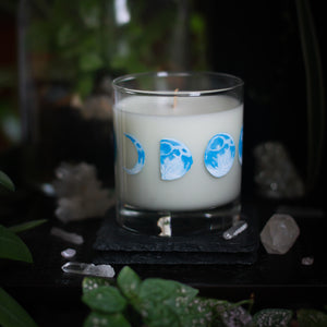 A white-wax candle with a wooden wick rests upon a dark surface with plants and crystals visible in the scene. The light candle stands out in contrast against the dark background. The candle glass has 8 phases of the moon painted in shades of blue circling the glass. This angle depicts the waxing half moon in the center with the waxing crescent moon to the left and the waxing gibbous moon on the right.