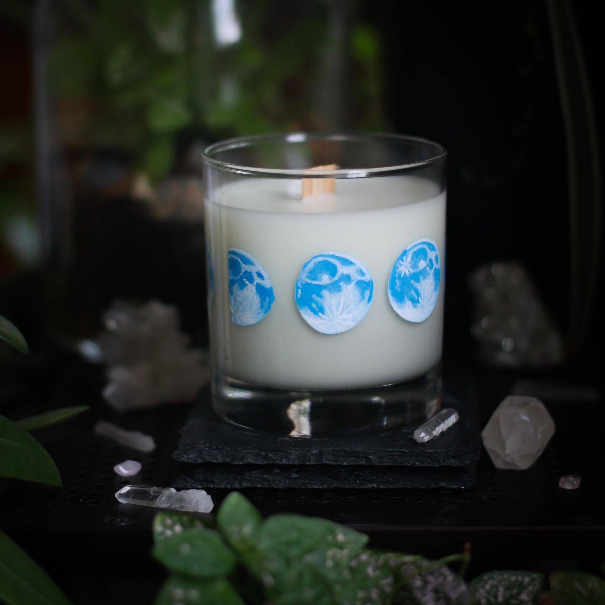 A white-wax candle with a wooden wick rests upon a dark surface with plants and crystals visible in the scene. The light candle stands out in contrast against the dark background. The candle glass has 8 phases of the moon painted in shades of blue circling the glass. This angle depicts the waxing gibbous moon in the center with the waxing half moon to the left and the full moon on the right.