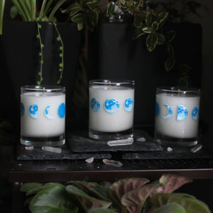 Three candle votives poured with white coconut-apricot wax each feature 8 hand-painted phases of the moon in shades of blue. The designs wraps around each glass.