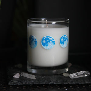 A candle votive poured with white coconut-apricot wax features 8 hand-painted phases of the moon in shades of blue. The design wraps around the glass.