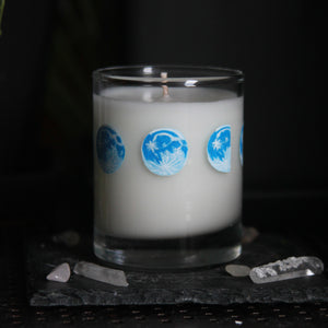 A candle votive poured with white coconut-apricot wax features 8 hand-painted phases of the moon in shades of blue. The design wraps around the glass.