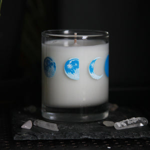 A candle votive poured with white coconut-apricot wax features 8 hand-painted phases of the moon in shades of blue. The design wraps around the glass. This angle shows the half and crescent moons.