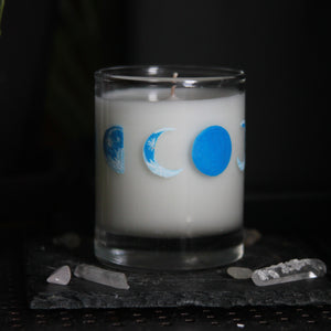 A candle votive poured with white coconut-apricot wax features 8 hand-painted phases of the moon in shades of blue. The design wraps around the glass. This angle displays the crescent and new moon.
