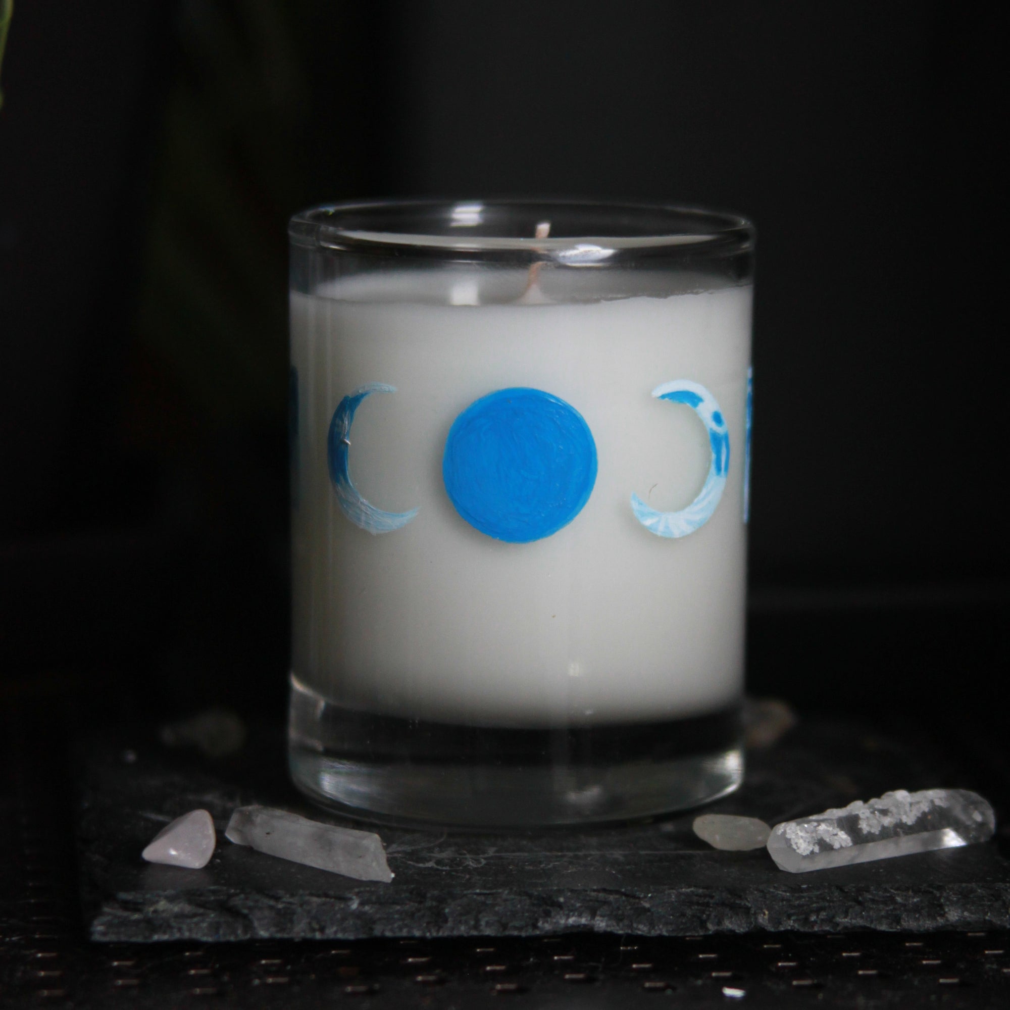 A candle votive poured with white coconut-apricot wax features 8 hand-painted phases of the moon in shades of blue. The design wraps around the glass. This angle shows a new moon painted all blue with crescent moons on either side facing it.