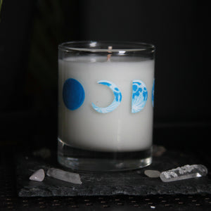 A candle votive poured with white coconut-apricot wax features 8 hand-painted phases of the moon in shades of blue. The design wraps around the glass.