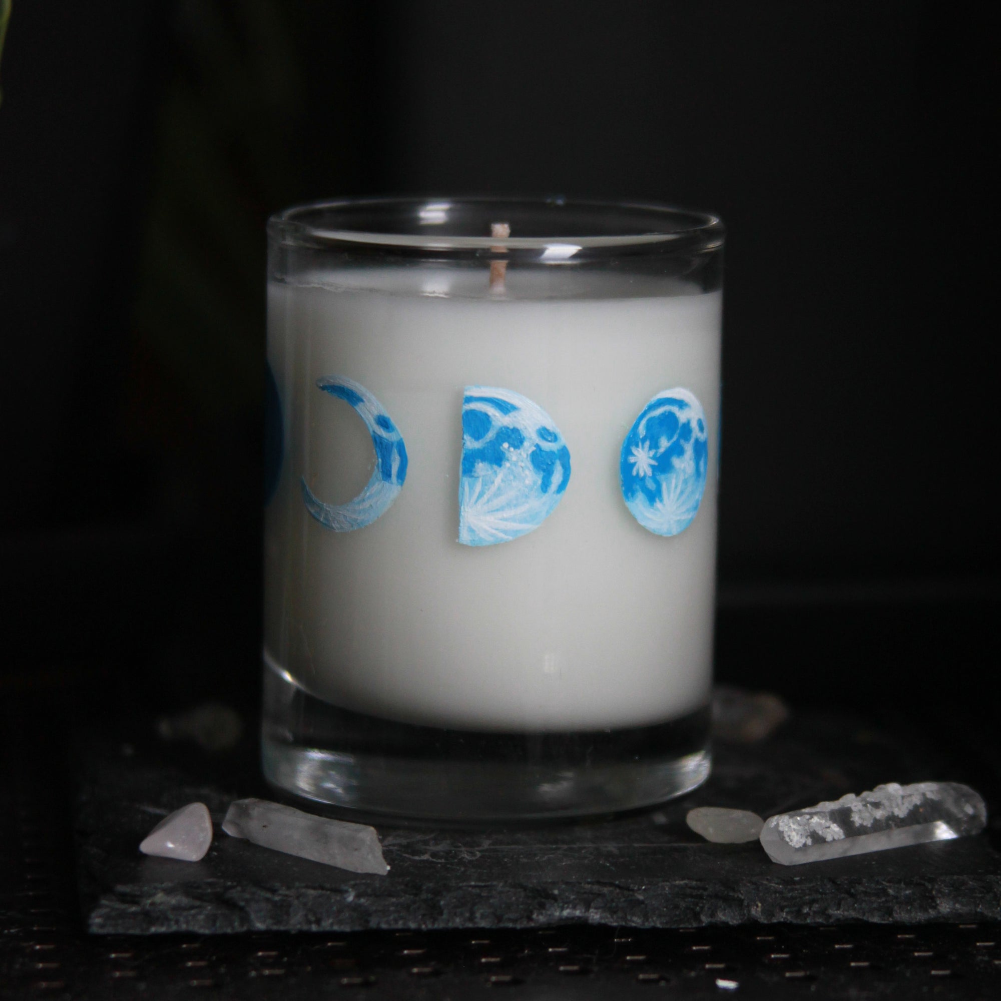 A candle votive poured with white coconut-apricot wax features 8 hand-painted phases of the moon in shades of blue. The design wraps around the glass.