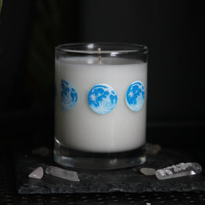A candle votive poured with white coconut-apricot wax features 8 hand-painted phases of the moon in shades of blue. The design wraps around the glass.