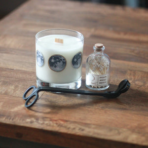 A white-wax candle with a wooden wick and a hand-painted moon phase design rests to the left of a small glass bottle with a cork top filled with wooden matches with white tips. In front of them lays a black matte wick trimmer. The candle has 8 phases of the moon painted in shades of gray with gold accents encircling the glass. The match bottle has a small white and gold label.