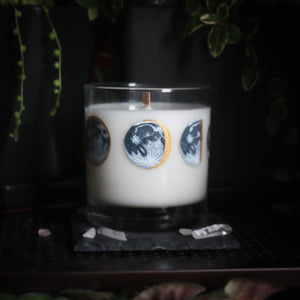 A white-wax candle with a wooden wick rests upon a dark surface with plants and crystals visible in the scene. The light candle stands out in contrast against the dark background. The candle glass has 8 phases of the moon painted in shades of gray with gold accents circling the glass. This angle depicts the waning gibbous moon centered with the full moon to the left and the waning half moon to the right.