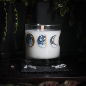 A white-wax candle with a wooden wick rests upon a dark surface with plants and crystals visible in the scene. The light candle stands out in contrast against the dark background. The candle glass has 8 phases of the moon painted in shades of gray with gold accents circling the glass. This angle depicts the waning half moon centered with the waning gibbous to the left and the waning crescent moon to the right