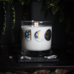 A white-wax candle with a wooden wick rests upon a dark surface with plants and crystals visible in the scene. The light candle stands out in contrast against the dark background. The candle glass has 8 phases of the moon painted in shades of gray with gold accents circling the glass. This angle depicts the waning crescent moon centered with the waning half moon to the left and the new moon to the right.