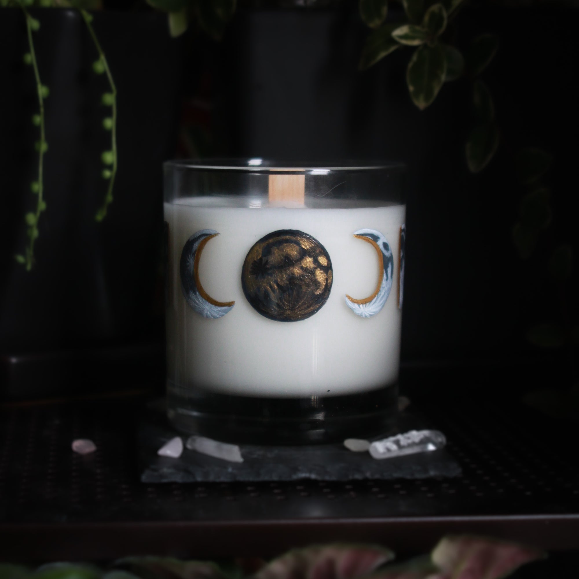A white-wax candle with a wooden wick rests upon a dark surface with plants and crystals visible in the scene. The light candle stands out in contrast against the dark background. The candle glass has 8 phases of the moon painted in shades of gray with gold accents circling the glass. This angle depicts the new moon centered with crescent moons on either side facing it. The new moon is painted in solid black with metallic gold shading.