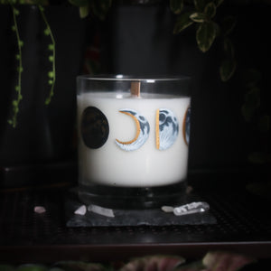 A white-wax candle with a wooden wick rests upon a dark surface with plants and crystals visible in the scene. The light candle stands out in contrast against the dark background. The candle glass has 8 phases of the moon painted in shades of gray with gold accents circling the glass. This angle depicts the waxing crescent moon centered with the new moon to the left and the waxing half moon to the right.