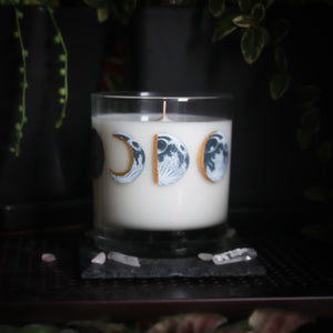 A white-wax candle with a wooden wick rests upon a dark surface with plants and crystals visible in the scene. The light candle stands out in contrast against the dark background. The candle glass has 8 phases of the moon painted in shades of gray with gold accents circling the glass. This angle depicts the waxing half moon centered with the waxing crescent moon to the left and the waxing gibbous moon to the right
