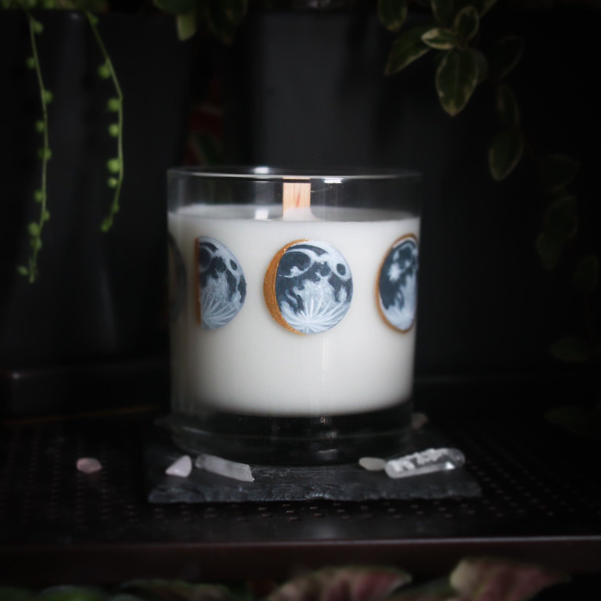 A white-wax candle with a wooden wick rests upon a dark surface with plants and crystals visible in the scene. The light candle stands out in contrast against the dark background. The candle glass has 8 phases of the moon painted in shades of gray with gold accents circling the glass. This angle depicts the waxing gibbous moon centered with the waxing half moon to the left and the full moon to the right.
